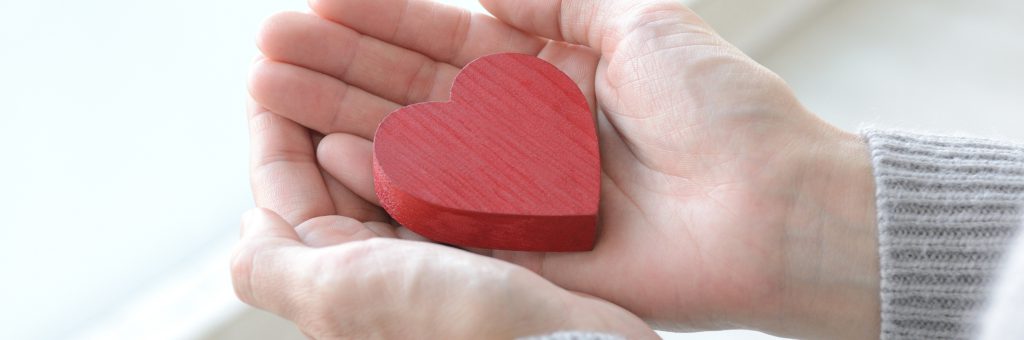 両手でハート型の木製オブジェクトを優しく包み込んでいるイメージ。支援・寄付活動を表す